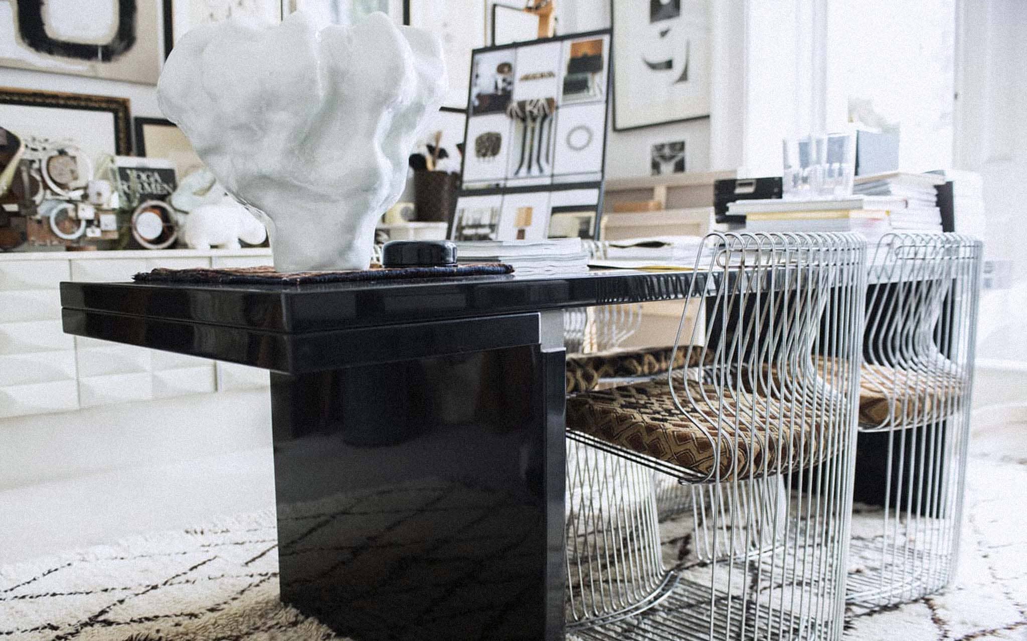 A large desk in glossy black with metal wire frame chairs. There is a rustic black and white patterned rug beneath the desk.There are various pieces of art and ceramic sculptures on display on the walls, and the desk. The colour palette is black, brown, white, and beige.